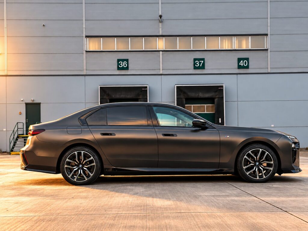 Matně šedé BMW i7 xDrive60 z pravého boku před halou při západu slunce